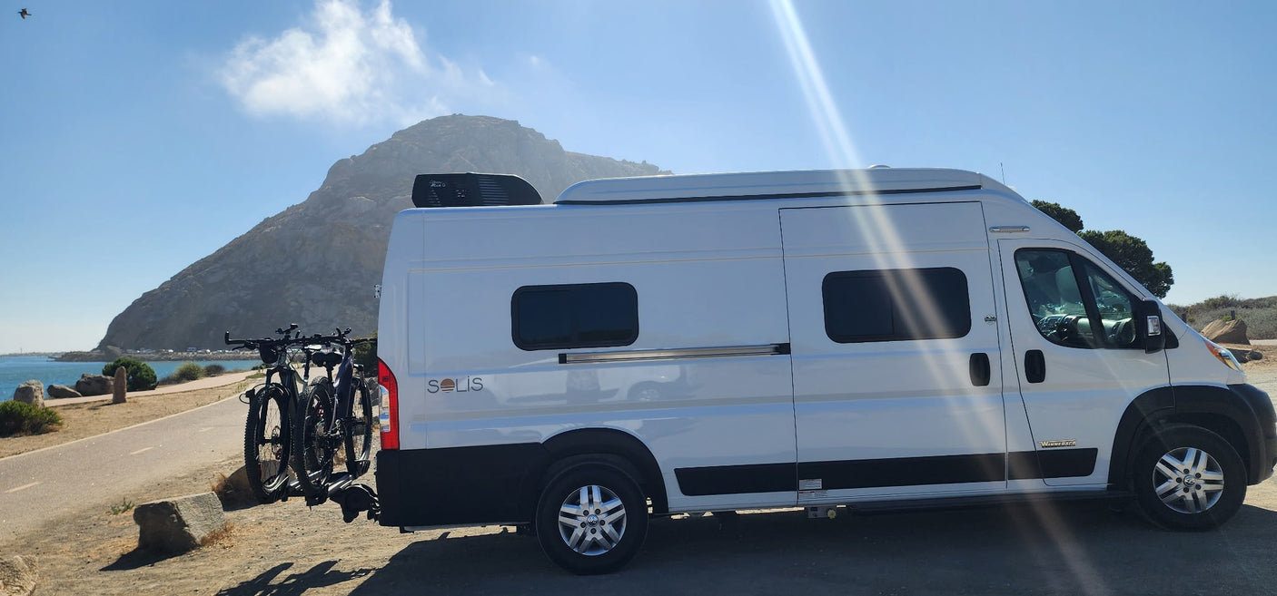 White Winnebago Solis RV rental with bikes by the sunny coast, a mountain in the background. Ready for adventure!