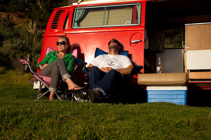 Two people relax outside their red RV rental, enjoying a sunny day of camping and leisure.