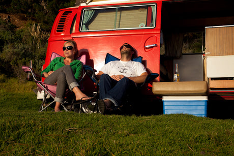 Two people relax outside their red RV rental, enjoying a sunny day of camping and leisure.