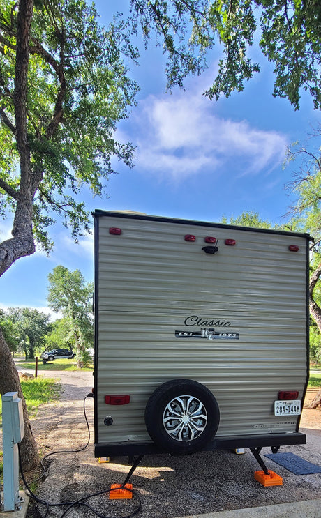 Rear view of an RV rental parked at a sunny campsite with trees, showing its spare tire and Texas license plate.