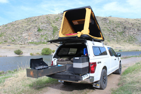 Overlanding truck with rooftop tent & outdoor kitchen by a scenic lake, perfect for an RV rental adventure.