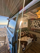 Beachfront RV rental: A blue camper van with open door revealing a cozy interior & ocean sunset views.
