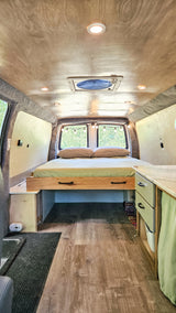 RV rental: Inside a cozy camper van featuring a rear bed, wood-look flooring, custom storage, and ceiling lighting.