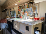 Well-equipped RV rental kitchen: counter, sink, microwave, and organized storage. Natural light brightens the cozy camper van interior.