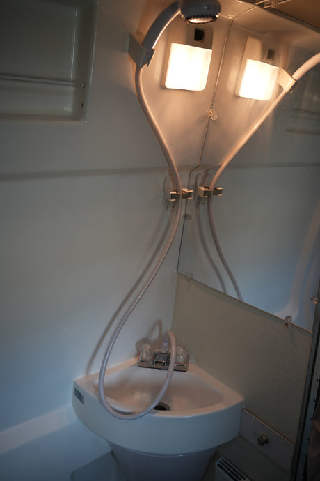 Inside an RV rental: a compact bathroom with a corner sink, dual-purpose shower head, mirror, and two bright overhead lights.