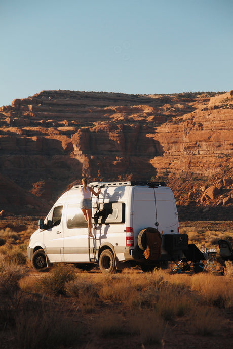 Woman climbing ladder on a white RV rental in a desert landscape with red cliffs. Remote van life adventure.