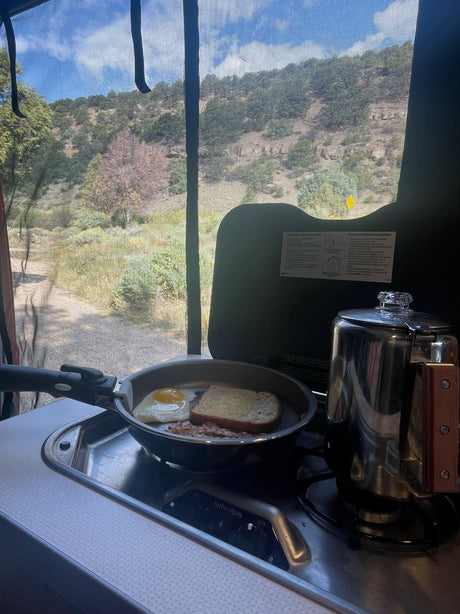 Inside an RV rental: cooking breakfast (egg, bacon, toast, coffee) on a compact stove, overlooking scenic hills & a path.
