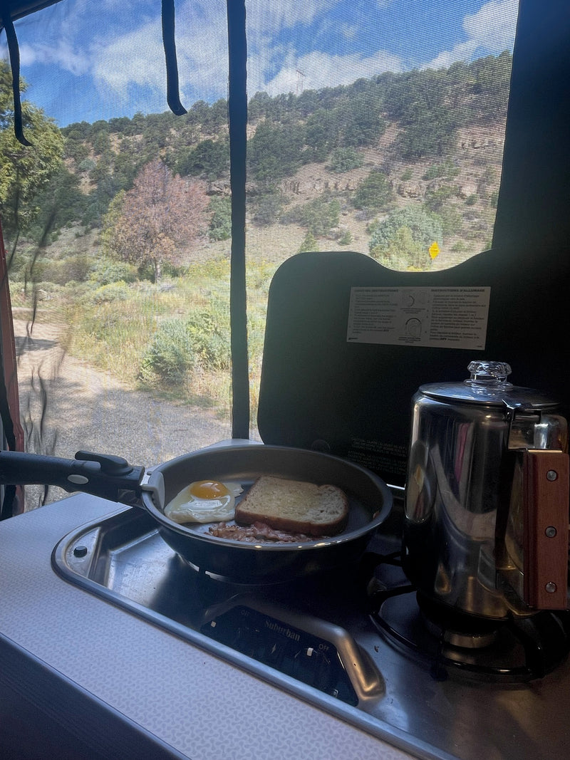 Inside an RV rental: cooking breakfast (egg, bacon, toast, coffee) on a compact stove, overlooking scenic hills & a path. — photo 11