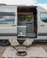 This RV rental shows an open camper van with an inviting outdoor setup, including two chairs, a fire pit, and a wine shelf.