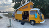 A man next to a yellow pop-top VW camper van on a sandy path by the water; ideal for rv rental travel.
