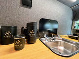 Inside an RV rental, a compact kitchen features a stainless steel sink, wood counter, and black Moxie Van Co. mugs.