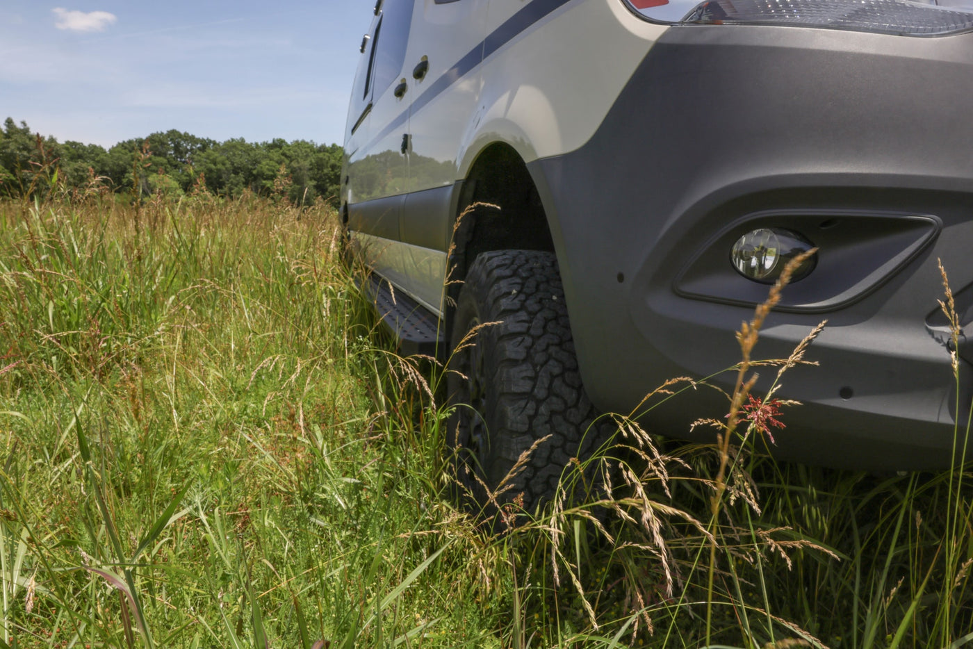 A low-angle view of an rv rental van's front right side, showing its off-road tire and fog light in tall, green grass. — photo 3