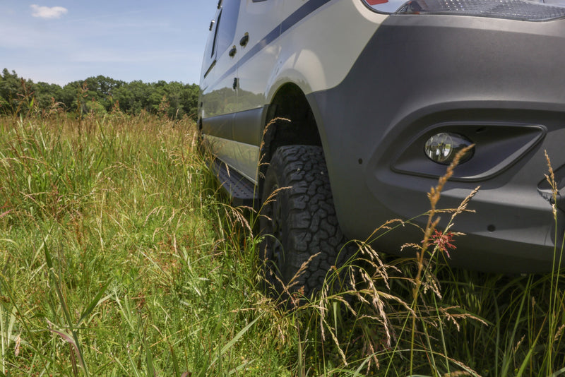 A low-angle view of an rv rental van's front right side, showing its off-road tire and fog light in tall, green grass. — photo 3