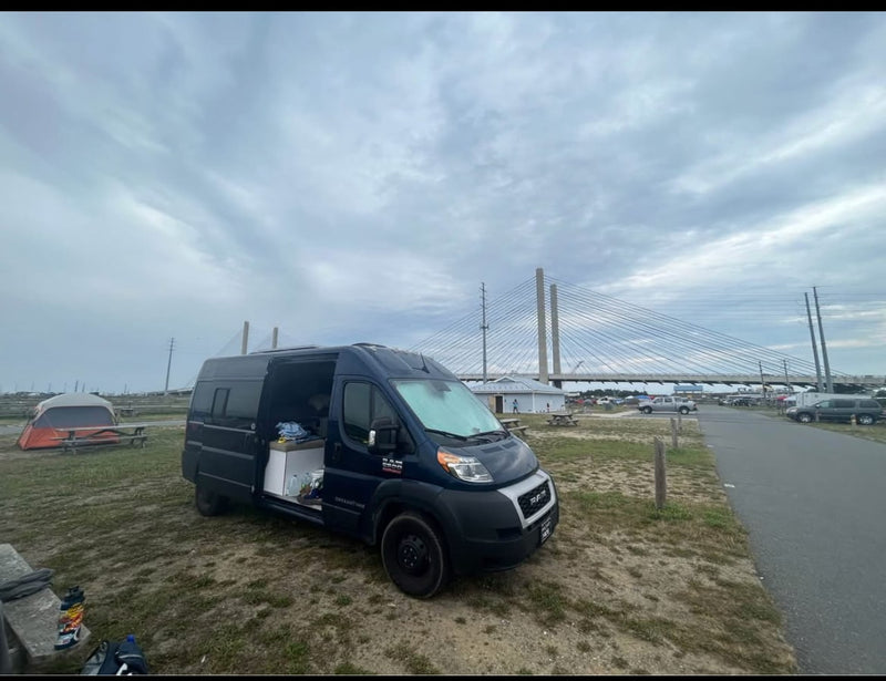 A blue camper van (RV rental) sits near a tent and a large cable-stayed bridge on a cloudy day, perfect for camping. — photo 3