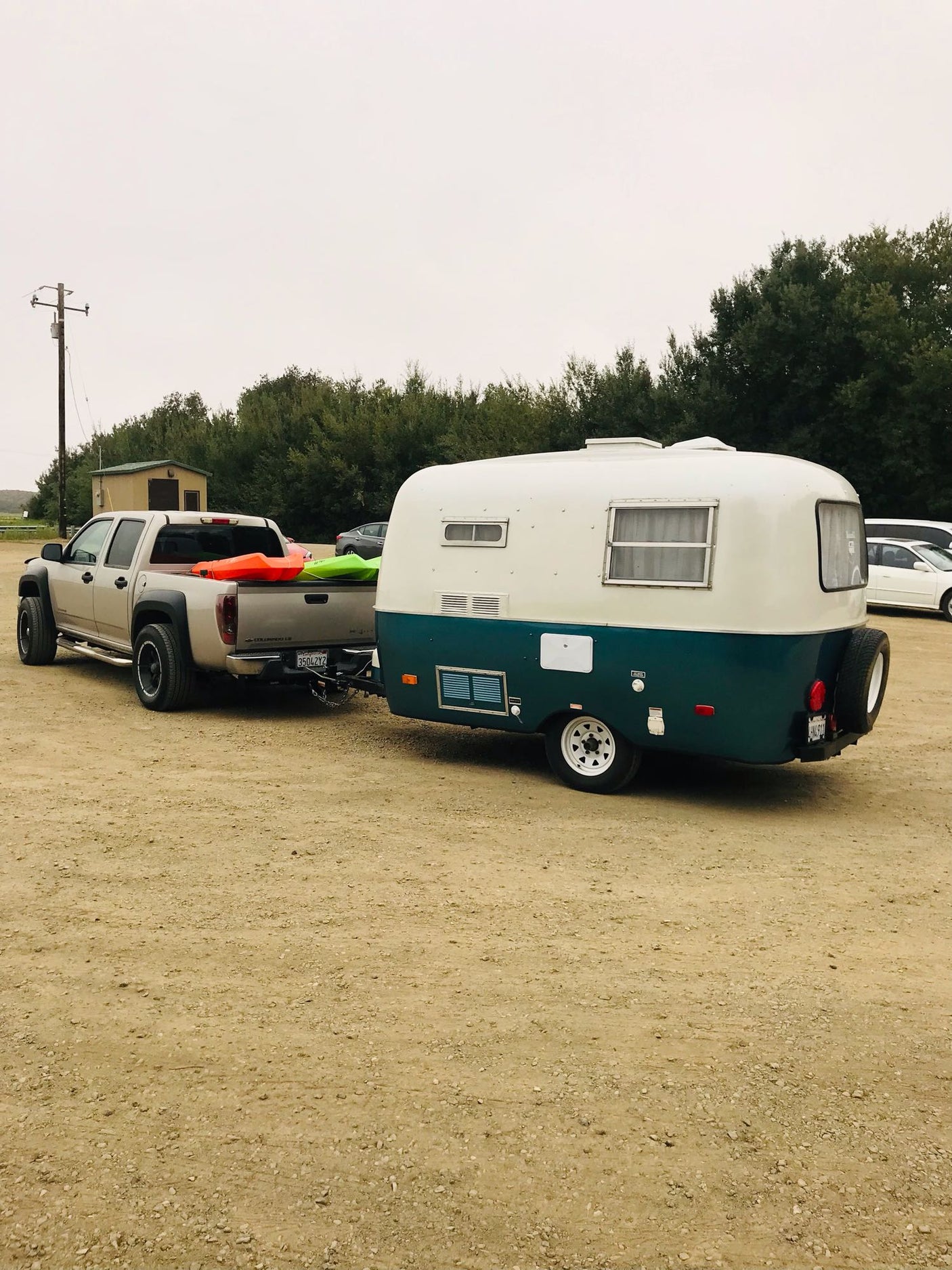 A pickup truck tows a white & teal retro RV rental, with kayaks loaded, parked on a dirt lot. Ready for adventure!
