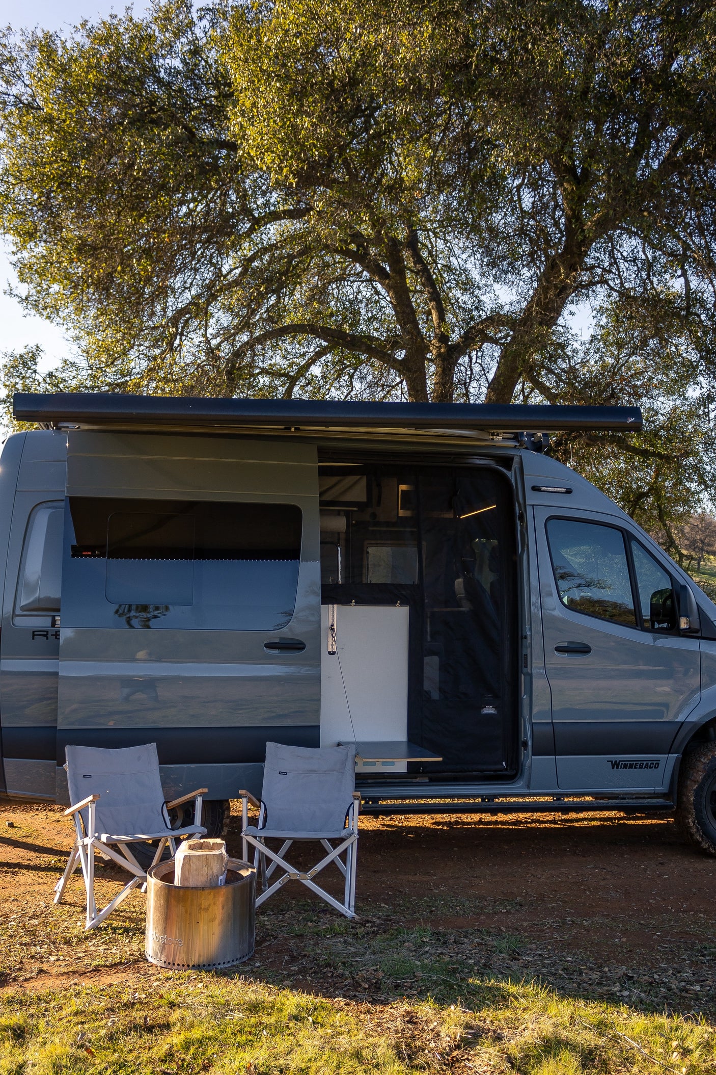 Grey camper van RV rental parked under a large tree, side door open, with camping chairs & fire pit set up for outdoor living.