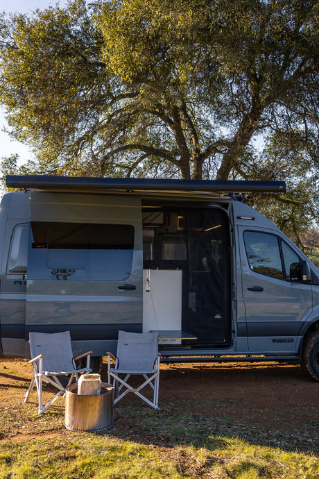 Grey camper van RV rental parked under a large tree, side door open, with camping chairs & fire pit set up for outdoor living.
