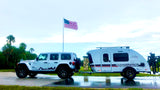 A white Jeep Wrangler tows a sleek Luna Rover RV rental on wet pavement, set against an American flag. Perfect for road trips.