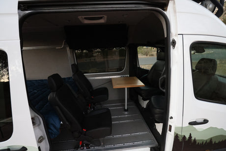 View inside a white RV rental: black modular seating, wood dining table, and a bed in the rear.