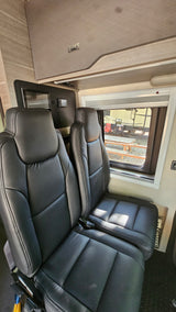 Inside a modern RV rental: Two black leather passenger seats, overhead storage, and window view of a campsite.