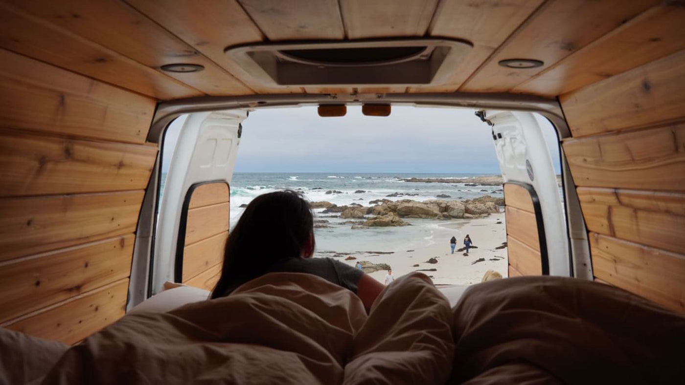 Wake up to ocean waves! This cozy **rv rental** shows a person on a bed inside a wood-paneled van, looking at a rocky beach. — photo 3
