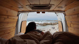 Wake up to ocean waves! This cozy **rv rental** shows a person on a bed inside a wood-paneled van, looking at a rocky beach.