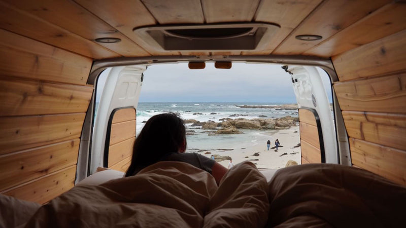 Wake up to ocean waves! This cozy **rv rental** shows a person on a bed inside a wood-paneled van, looking at a rocky beach. — photo 3