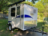 A white compact Runaway Rouser RV rental teardrop camper, featuring an awning, AC, and front cargo rack, parked on asphalt.