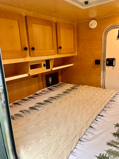 Cozy RV rental interior featuring a comfortable bed with a cable-knit blanket, wooden cabinets, and storage.