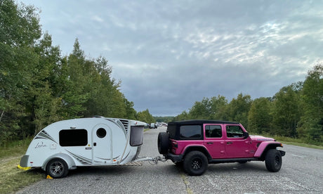 Pink Jeep Rubicon towing a white Luna teardrop RV rental on a paved road, surrounded by trees. Perfect for camping!