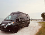 A dark grey Ram ProMaster camper van is parked on a dirt road next to water, ideal for an rv rental adventure.