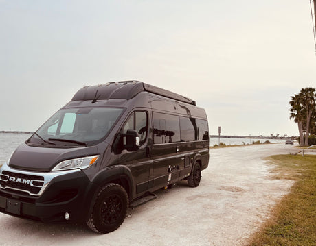 A dark grey Ram ProMaster camper van is parked on a dirt road next to water, ideal for an rv rental adventure.
