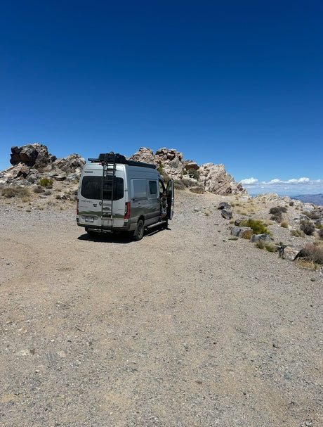 Grey RV rental camper van parked on a gravel path in a rocky desert, side door open under a vast blue sky. Adventure travel.