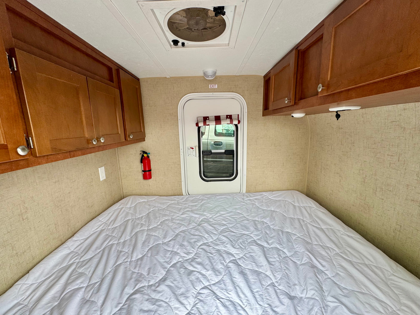 Cozy RV rental interior: large bed, wooden cabinets, ceiling fan, exit door, and fire extinguisher for safety.