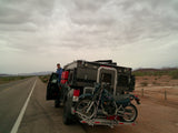 Adventure-ready RV rental: Truck camper with motorcycle carrier parked roadside in desert landscape. A person stands by the driver's door.