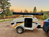 White teardrop camper RV rental with a side awning, hitched to a car in a scenic, tree-filled landscape at sunset.