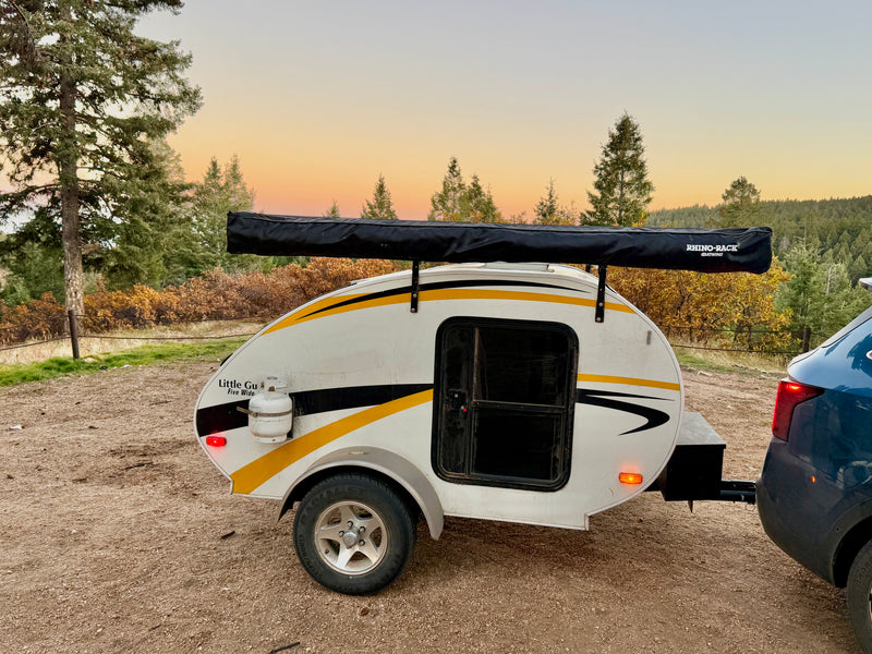 White teardrop camper RV rental with a side awning, hitched to a car in a scenic, tree-filled landscape at sunset. — photo 1