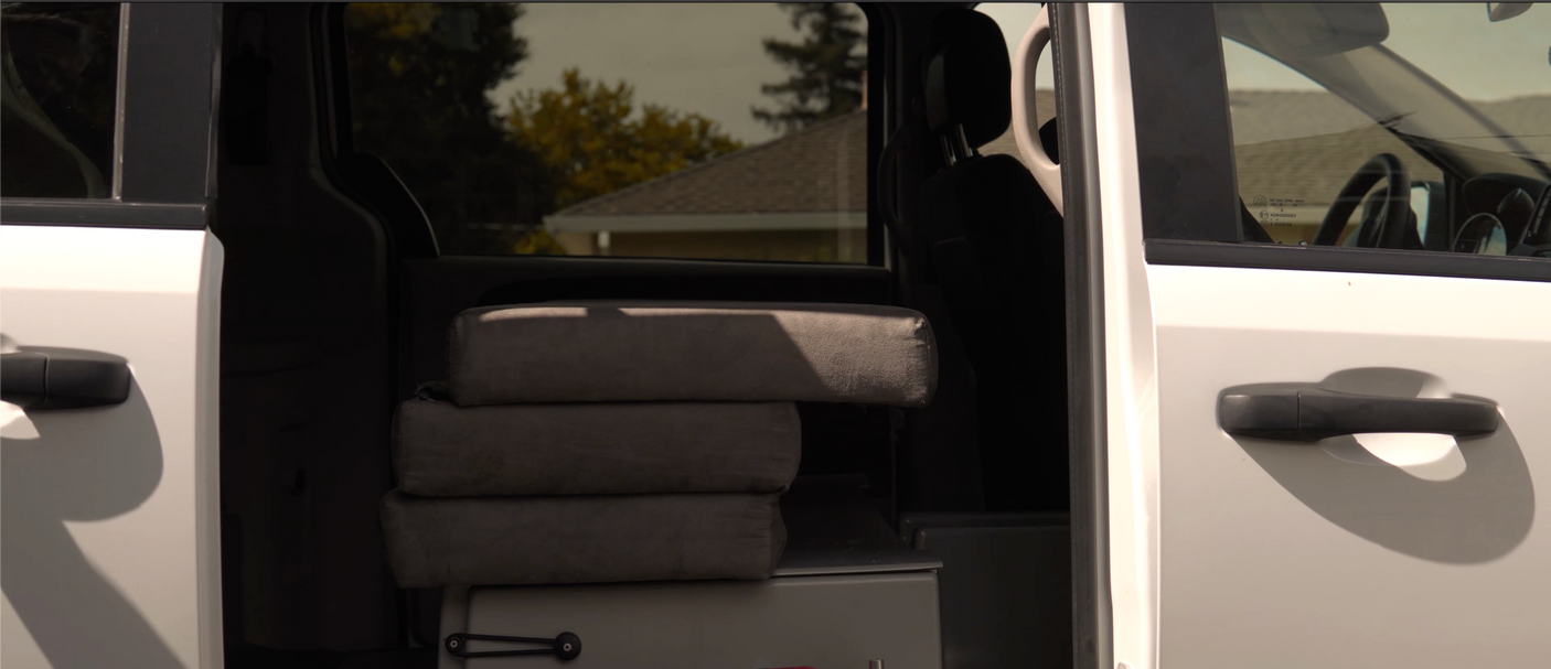 Inside an RV rental: three stacked gray cushions, likely a bed, visible through the open side door of the white van.