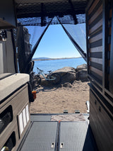 Inside an RV rental: Looking out at a mountain bike on sandy shore by a lake under clear blue sky.