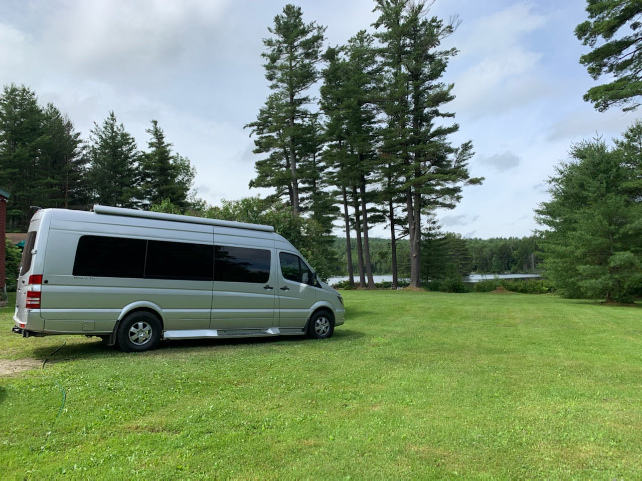 Silver RV rental camper van parked on a green lawn by a scenic lake, surrounded by tall pine trees under a cloudy sky.