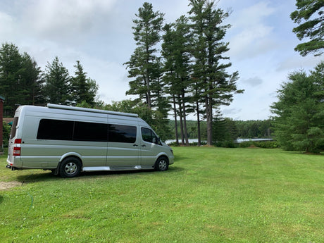 Silver RV rental camper van parked on a green lawn by a scenic lake, surrounded by tall pine trees under a cloudy sky.