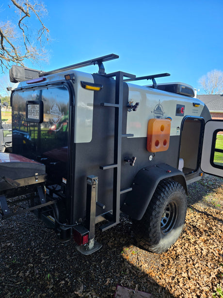 This compact off-road travel trailer, ideal for an RV rental, features robust tires, a roof rack, and side ladder.