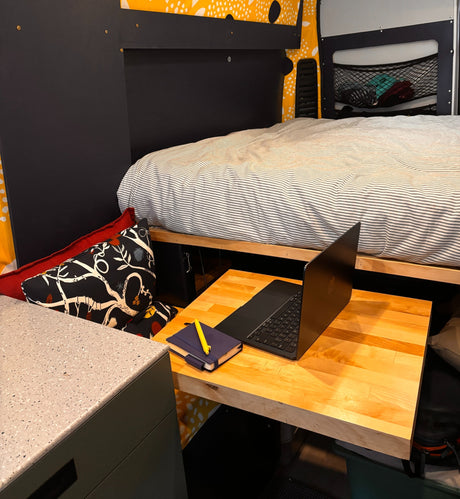 RV rental interior features a striped bed, wooden desk with laptop, and cushion. Compact workspace for travel.