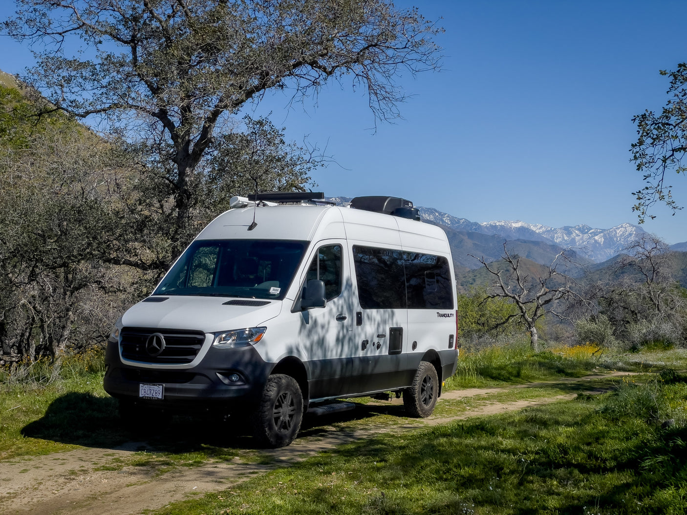 White Winnebago Tranquility Sprinter RV rental on a dirt path with trees and snowy mountains under a clear sky.