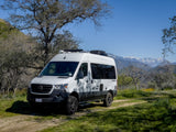 White Winnebago Tranquility Sprinter RV rental on a dirt path with trees and snowy mountains under a clear sky.