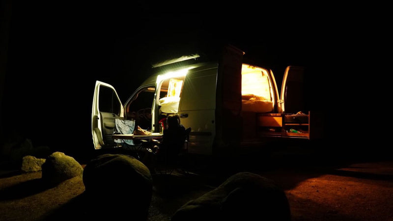 Night scene: a bright RV rental with open doors reveals a cozy interior. A person sits outside at a table near rocks. — photo 4