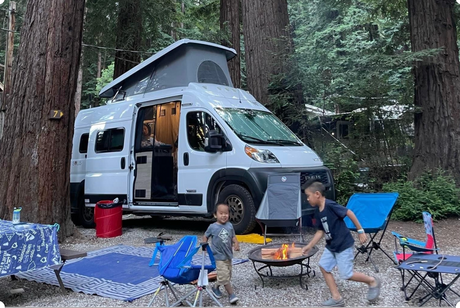 Family fun at a redwood campsite with their rv rental. Kids play by the fire, campervan door open, enjoying nature.