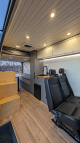 Spacious RV rental interior: wood floor, modern kitchen, passenger seats, dinette, and a comfy elevated bed.