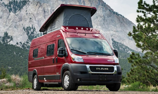 Red pop-top camper van for RV rental, parked on a dirt path with rugged mountains in the background.