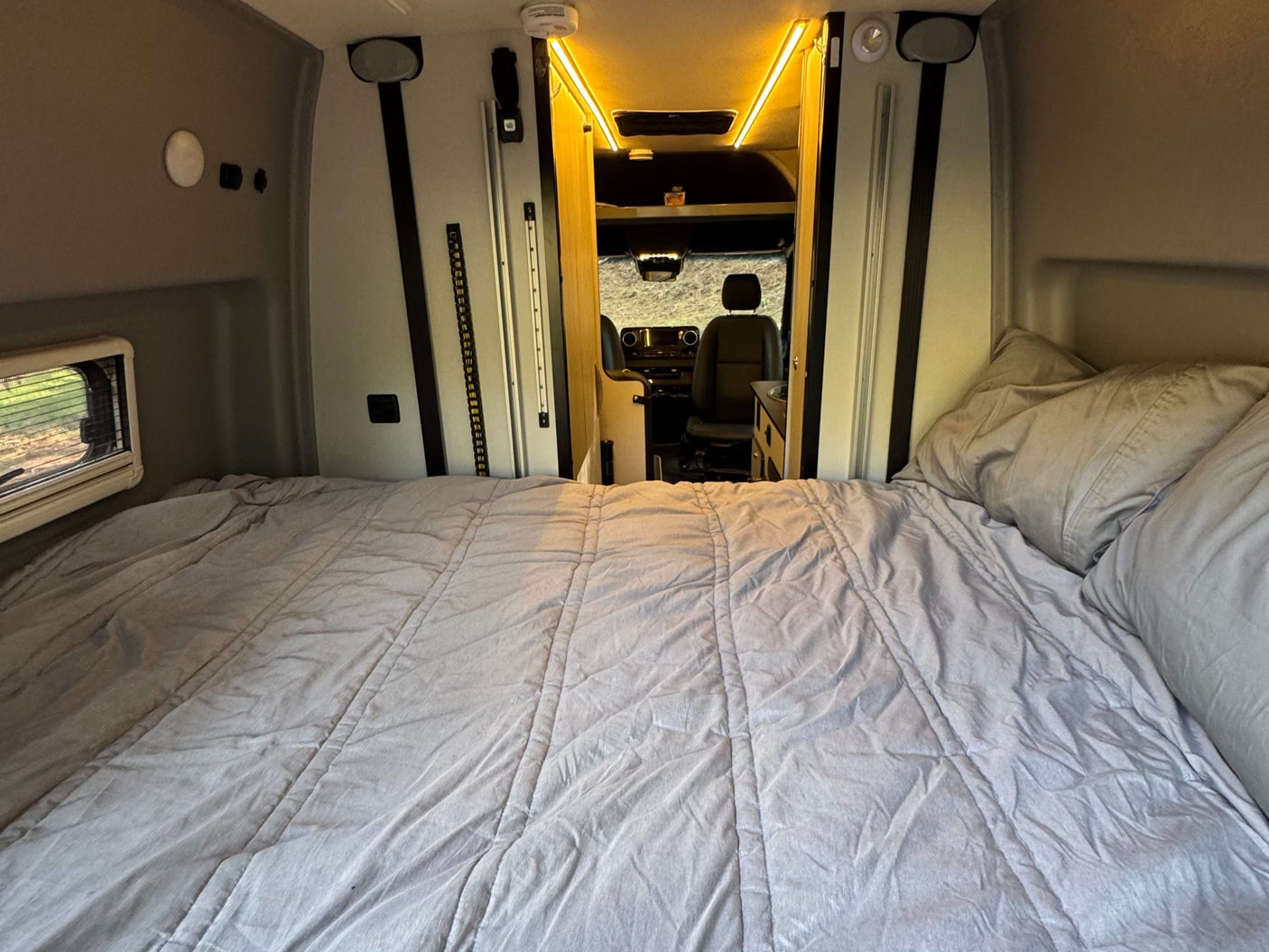 Cozy RV rental interior with a bed in the foreground and the driver's cabin visible. Bright lights illuminate the ceiling.